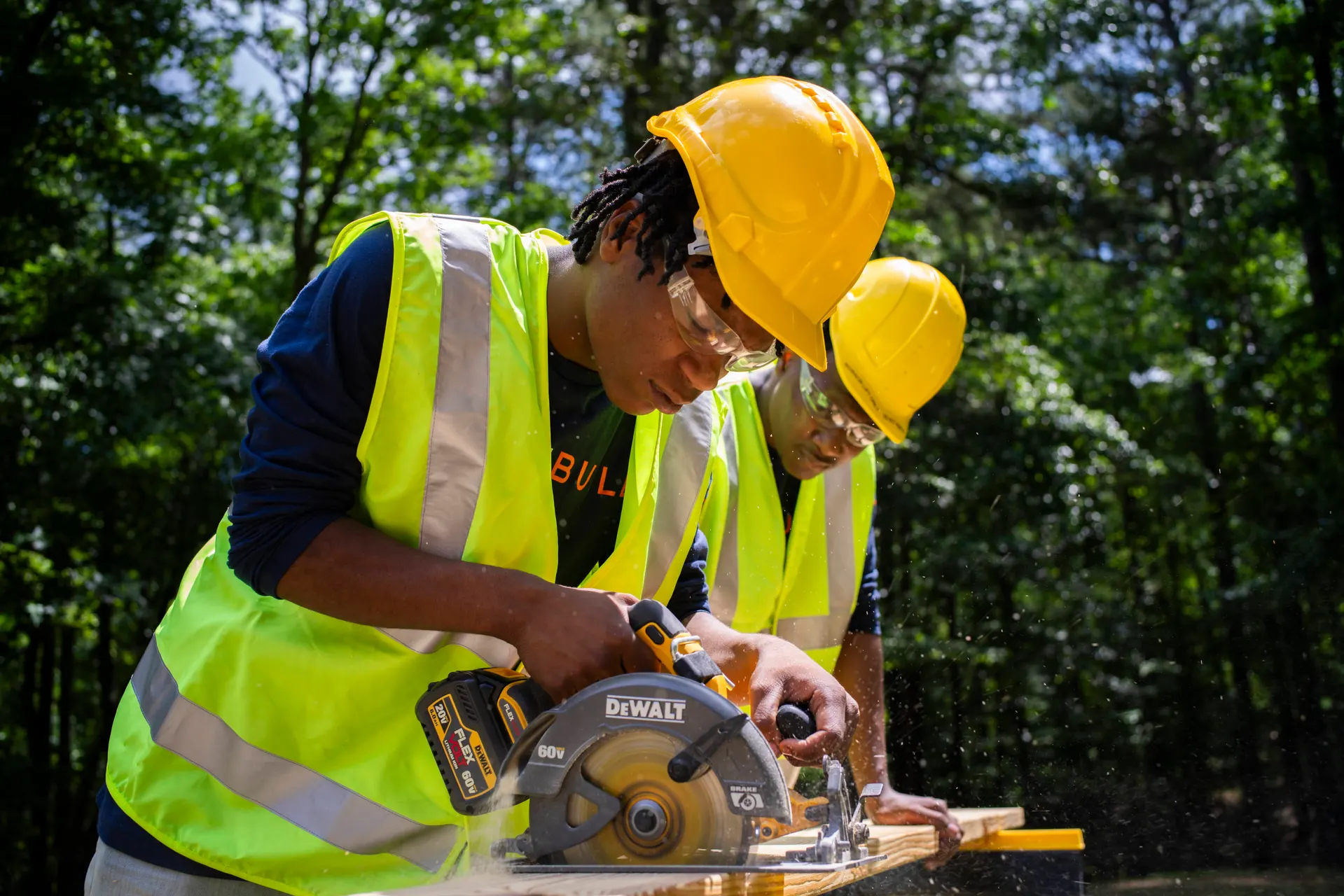 Two YouthBuild participants in hard hats cutting lumber with a circular saw