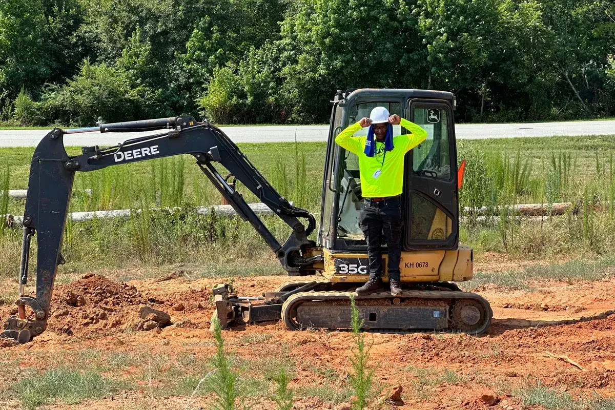 A participant in the cab of a mini excavator with both arms raised