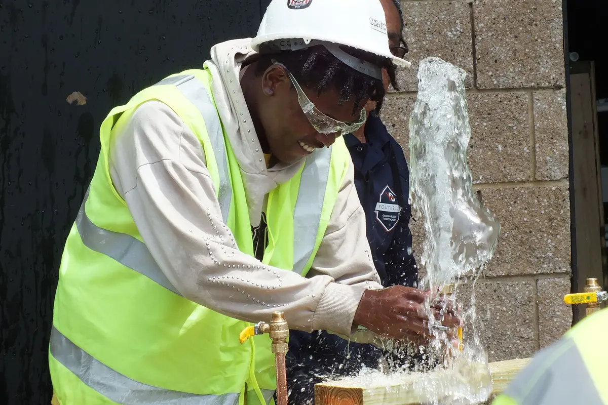 A participant in safety glasses grins as water sprays from a fitting on the plumbing bench