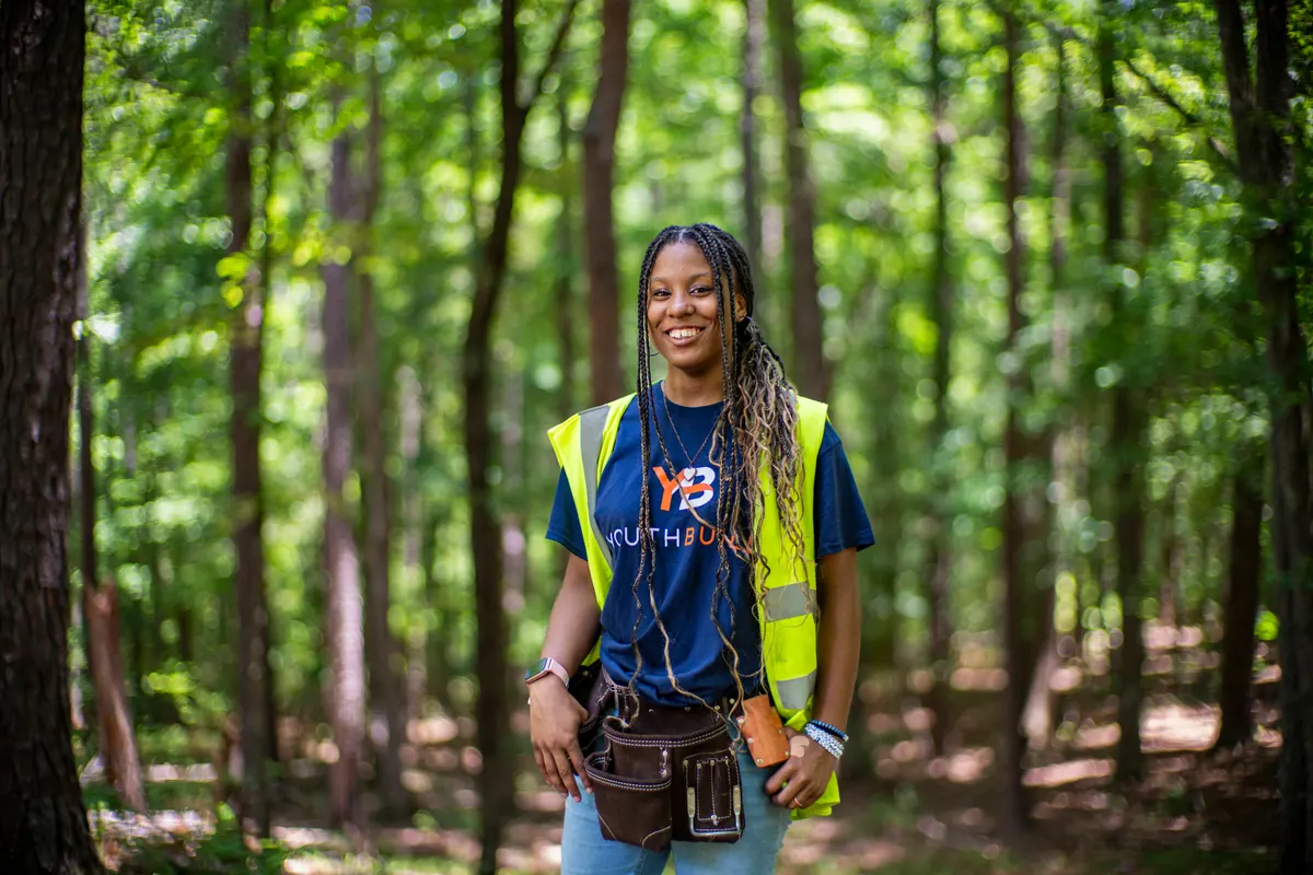 A smiling YouthBuild participant in a hi-vis vest and tool belt in the woods